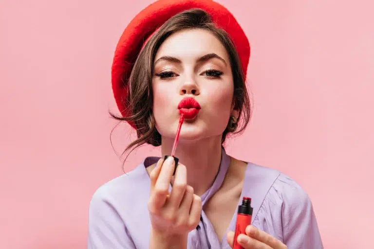 Femme en chemisier coiffée d’un béret rouge se maquillant avec un gloss longue tenue