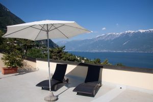Parasol blanc sur une terrasse protégeant deux chaises longues du soleil, avec un lac et une chaîne de montagnes