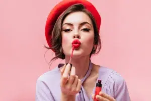 Femme en chemisier coiffée d’un béret rouge se maquillant avec un gloss longue tenue