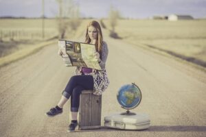 Femme assise au milieu d'une route déserte qui regarde une carte routière à côté d'une valise et d'un globe terrestre
